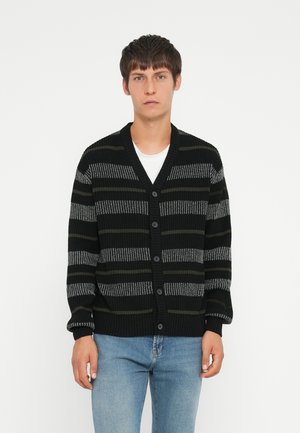 Schwarz-grau gestreifter Cardigan mit V-Ausschnitt, Knopfschluss und gerippten Bündchen. Getragen über einem weißen T-Shirt und kombiniert mit blauen Jeans.