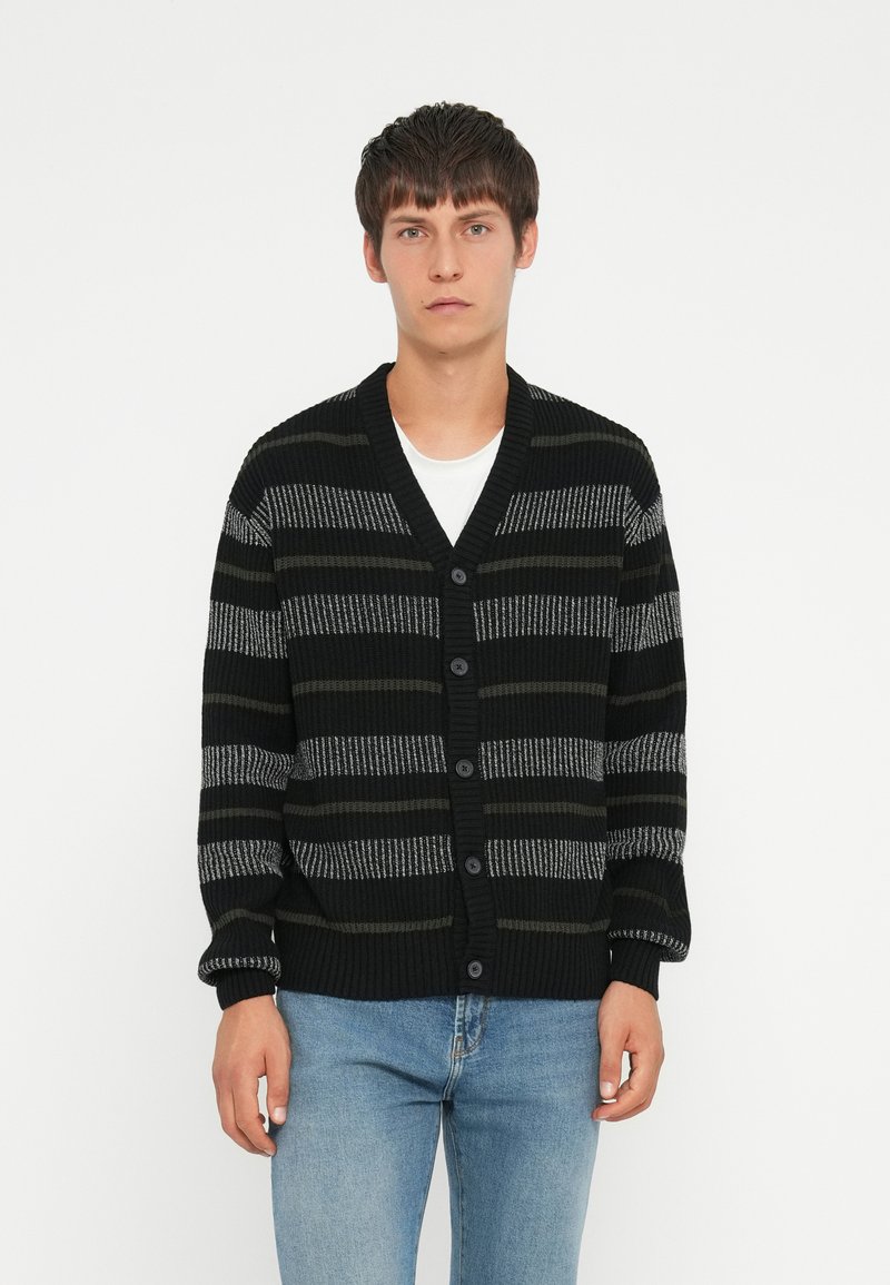 Black and grey striped cardigan with a v-neck, button closure, and ribbed cuffs. Worn over a white t-shirt and paired with blue jeans.