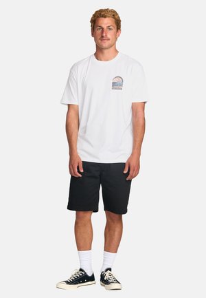 Hvid bomulds t-shirt med et grafisk motiv af palmer, parret med sorte shorts og sorte sneakers. Enkle hvide sokker er synlige.