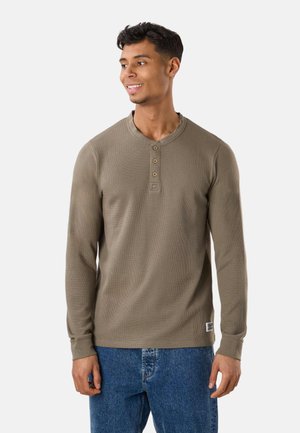 Mężczyzna w brązowej koszulce henley z długim rękawem i niebieskich dżinsach, stoi i uśmiecha się, patrząc w lewo.