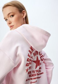 Sudadera de color rosa claro hecha de una mezcla de algodón, con un gran gráfico rojo en la parte posterior que incluye texto y una estrella. Manga larga, corte relajado y capucha.