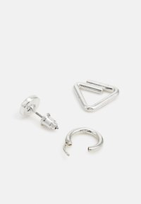 Três brincos metálicos prateados: um stud com uma base redonda, um argola com um design aberto e um em forma triangular. Textura suave, com detalhes mínimos.