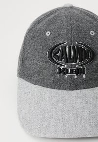 Grå filthat med en mørkere grå øverste sektion, lys grå skygge, med et fremtrædende sort broderet "CALVIN KLEIN" logo og 1968 detalje.