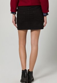 Mustahidis musta denimi mini etek, düz kesim, arka cepler ve temiz bir etek ucu ile, siyah bilek botlarıyla kombinlenmiş.