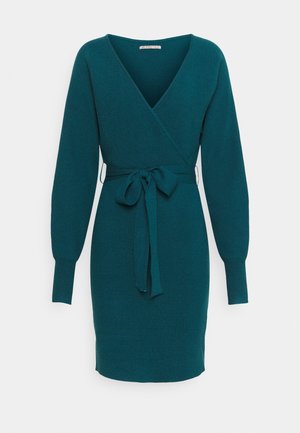 Robe enveloppante tricotée en bleu sarcelle avec des manches longues, un décolleté en V et une taille nouée. Texture douce, forme ajustée et longueur au milieu de la cuisse.