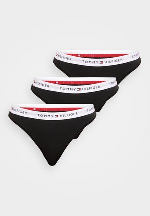 Trīs melnas sieviešu biksītes ar baltām elastīgām lentēm, uz kurām rakstīts "Tommy Hilfiger", un sarkanu iekšpusi, sakārtotas pārklājoties uz baltas virsmas.