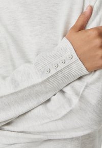 Light gray ribbed sweater sleeve with four small white buttons near the wrist, hand resting on opposite arm.