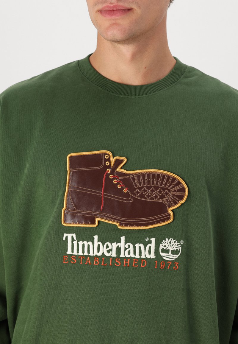 Uomo che indossa una felpa verde con una grafica di stivali marroni e il testo "Timberland Established 1973" sul petto.