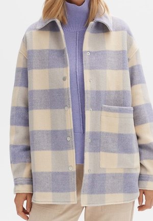 Frau trägt eine creme- und lavendelfarbene karierte Jacke mit Knöpfen über einem lavendelfarbenen Rollkragenpullover und beigen Hosen.
