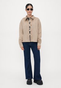 Beige suede jacket with a collared neckline and wooden buttons, layered over a patterned top. The model wears dark flared jeans and chunky black shoes.