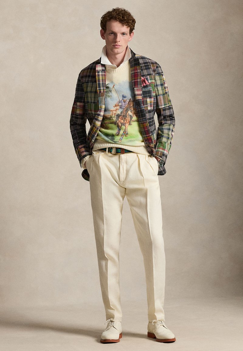 Giovane uomo che indossa un blazer a quadri multicolore, un maglione con grafica di un giocatore di polo, pantaloni crema plissettati e scarpe eleganti bianche, posando con le mani nelle tasche.