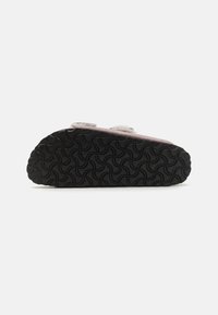 Roze slipper met een gestructureerde zwarte rubberen zool, voorzien van een golfpatroon. Klittenbandsluiting aan de bovenkant zorgt voor gemakkelijk aan- en uittrekken. Gemaakt van zachte materialen.