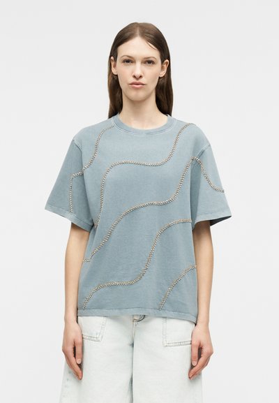 Νεαρή γυναίκα με ίσια καστανά μαλλιά φοράει ένα φαρδύ μπλε μπλουζάκι με κυματιστά σχέδια από στρας και ανοιχτόχρωμα τζιν με φαρδιά μπατζάκια.