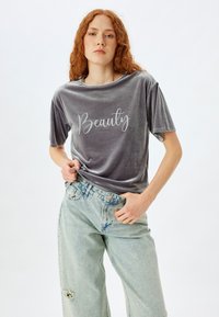 Camiseta de terciopelo gris con mangas cortas que presenta la palabra "Belleza" en letras cursivas blancas, combinada con jeans de mezclilla claros y desgastados.