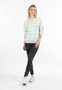 Helleblau gestrickter Pullover im Hahnentrittmuster mit Rundhalsausschnitt und gerippten Bündchen, kombiniert mit dunklen Jeans und weißen Sneakers.