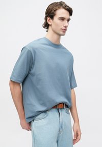 Jauns vīrietis ar brūniem matiem, ģērbies brīvā piegriezuma zila īsām piedurknēm kreklā, kas ielikts gaiši zilās džinsās ar brūnu jostu, uz balta fona.