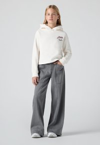 Jaunas žmogus stovi, apsirengęs baltu „Levi's“ logotipo hoodie, laisvomis pilkomis kelnėmis ir baltais sportbačiais prieš paprastą šviesią fono.