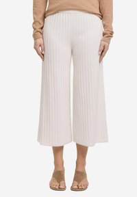 Białe spodnie typu wide-leg z prążkowaną teksturą, o luźnym kroju i elastycznym pasie. Stylizowane z brązowymi klapkami.