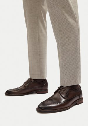 Braune Brogue-Lederschuhe mit perforierten Details, kombiniert mit hell karierten Hosen. Mit abgerundetem Zehenbereich und geschnürtem Vorderteil.