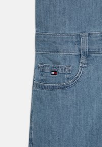 Světlé modré džínové kalhoty s malým vyšívaným logem v červené, bílé a modré barvě na zadní kapse. Textura je hladká s viditelným šitím.