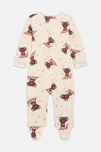 Κρεμ φορμάκι μωρού με μακριά μανίκια και καλυμμένα ποδαράκια, με επαναλαμβανόμενο μοτίβο αρκουδάκι που φοράει κόκκινο πουκάμισο και μπλε παντελόνι με το κείμενο "Guess".