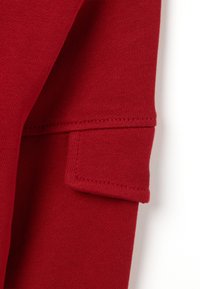 Tejido de algodón rojo con una textura suave, que presenta un sutil patrón acanalado y un detalle de bolsillo cosido en la manga.