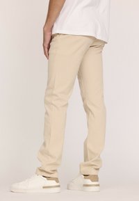 Chinos beiges avec une coupe slim, fabriqués en tissu coton, dotés de poches latérales et d'un léger effilément vers les chevilles, associés à des baskets blanches.