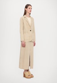 Beige denimblazer med en knapp, två framfickor, lager på lager över en matchande midi-kjol med en framfåra, parat med beige plattformssandaler.