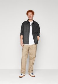 Sort tekstureret skjorte over hvid T-shirt, parret med beige cargo-bukser. Hvide sko med marineblå snørebånd og beige såler. Enkel, afslappet stil.