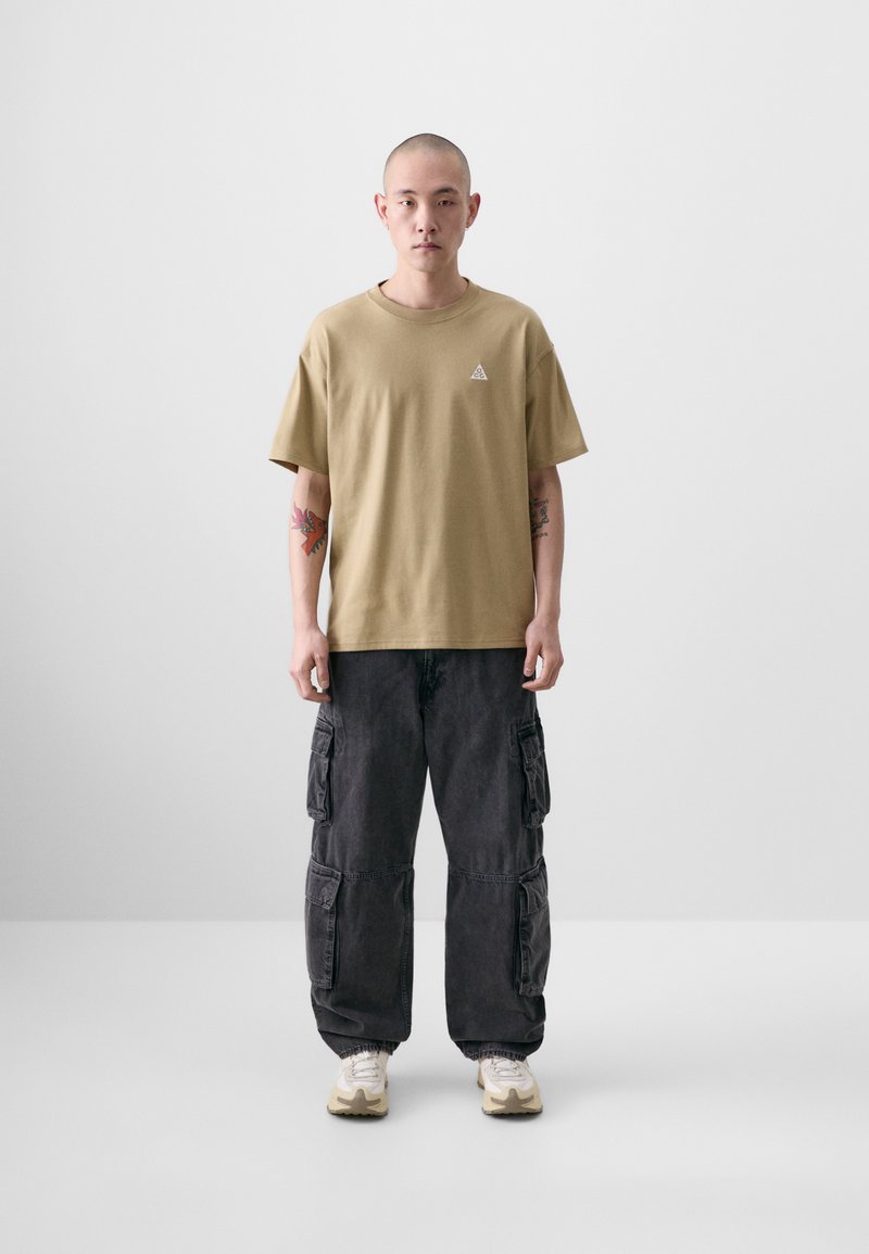 Oliven grøn kortærmet t-shirt med et lille trekantet logo på brystet, parret med mørkegrå cargobukser med flere lommer.