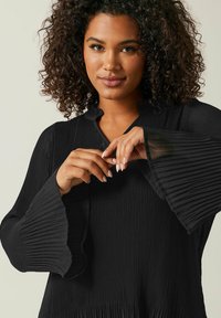 Schwarze plissierte Bluse mit hohem Kragen und Glockenärmeln. Der strukturierte Stoff erzeugt eine fließende Silhouette mit einem sanften, leichten Aussehen.