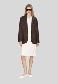 Blazer en daim marron sur une robe blanche, assorti à des ballerines blanches. Les accessoires incluent des lunettes de soleil et un collier de perles. Design simple et moderne.