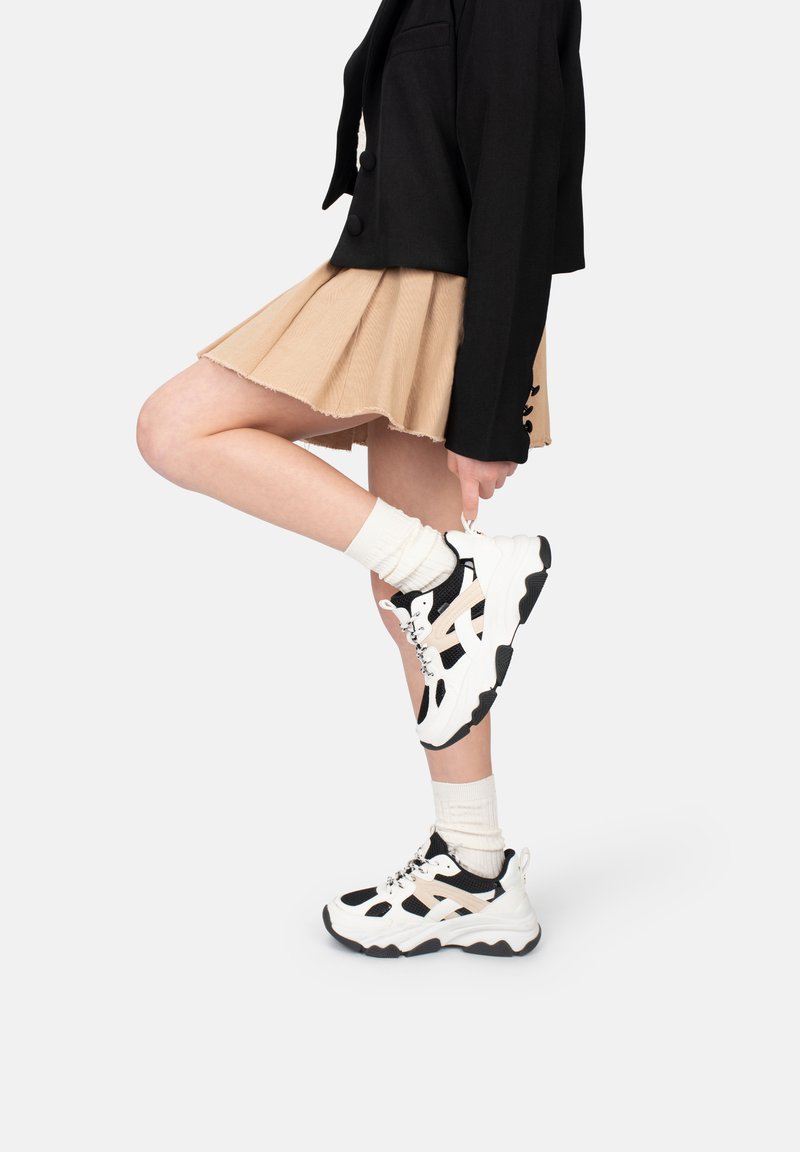 Schwarze und beigefarbene Sneaker aus Mesh und strukturiertem Stoff mit einer klobigen Sohle, weißen Schnürsenkeln und einem schwarzen Akzentmuster.