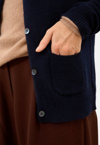 Marinblå ullkofta med ribbstickade ärmmuddar, med en framficka och rund knappdetalj. Lagd över en beige topp och bruna byxor.