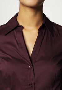 Camisa burdeos de cuello en tejido suave, con un delantero abotonado y detalles de costura visibles, de diseño ajustado y ligero brillo.