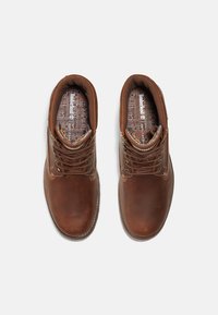 Botas de cuero marrón hasta el tobillo con puntera redonda, cuello acolchado, costuras en contraste y ojetes metálicos para los cordones. Diseño de plantilla texturizada visible.