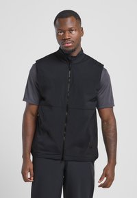 Schwarze ärmellose Weste mit hohem Kragen, Reißverschluss vorne und zwei Seitentaschen, aus leichtem, glattem Stoff, kombiniert mit einem dunkelgrauen Shirt.