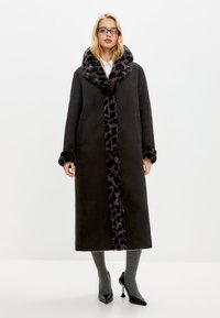 Lange zwarte jas met een leopardprint bontkraag en -accenten, met zijzakken en een rechte snit, gedragen over grijze sokken en zwarte hakken.