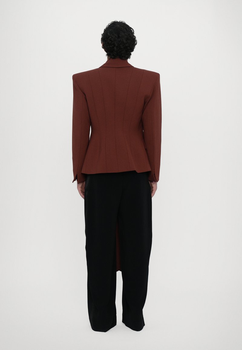 Brauner strukturierter Blazer mit vertikalen Nähten, Reverskragen und langen schwarzen Hosen mit einem Schlitz hinten. Glatte Stofftextur.