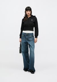 Sudadera negra recortada con cremallera frontal, combinada con vaqueros de pierna ancha de color azul claro que cuentan con dos bolsillos delanteros y herrajes de tono dorado.