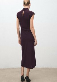Mujer con el pelo recogido, vistiendo un vestido midi sin mangas de color púrpura oscuro y tacones negros, de pie contra una pared blanca lisa.