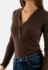 Haut marron côtelé à manches longues avec un décolleté en V plongeant et trois boutons au niveau du col, associé à un jean en denim bleu avec un bouton en argent.