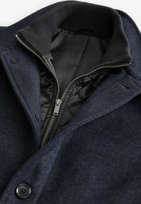 Marineblaue Fischgrätenjacke mit einem schwarz gefütterten Reißverschluss, geripptem Kragen und Knopfverschluss; eleganter Textur und strukturiertem Design.