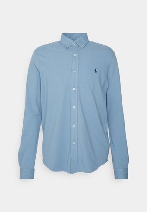 Chemise bleu clair à manches longues boutonnée avec col et petit logo sombre visible sur le côté gauche de la poitrine.
