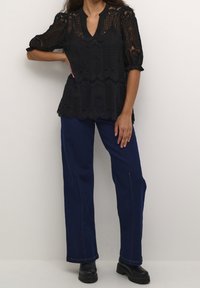 Blusa de encaje negro con escote en v y mangas de largo medio, diseño en capas, combinada con pantalones de talle alto de color azul oscuro y botas negras gruesas.