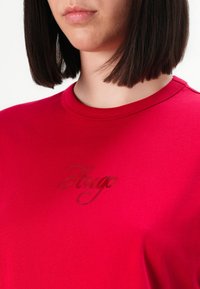 Donna con capelli scuri e lisci che indossa una camicia rossa con la scritta "Hugo" ricamata in corsivo sul petto.