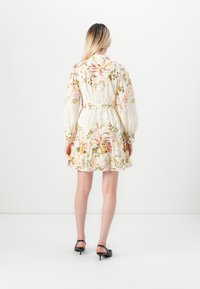 Donna con lunghi capelli biondi che indossa un vestito floreale con cintura e orlo arricciato, con tacchi neri, vista da dietro su uno sfondo bianco.