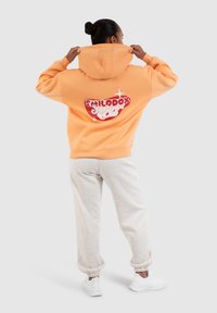 Haut à capuche orange avec un logo graphique au dos, associé à un pantalon de survêtement gris clair. Le matériau semble doux et décontracté.