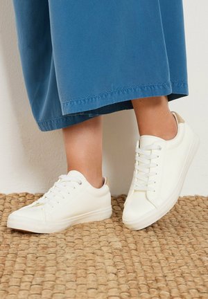 Person wearing white low-top sneakers with laces and blue wide-leg pants on a textured beige rug.