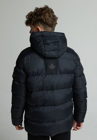 Doudoune noire avec capuche, présentant un design matelassé, des poignets élastiques et un logo de feuille d'érable noire sur le panneau arrière. Texture lisse.